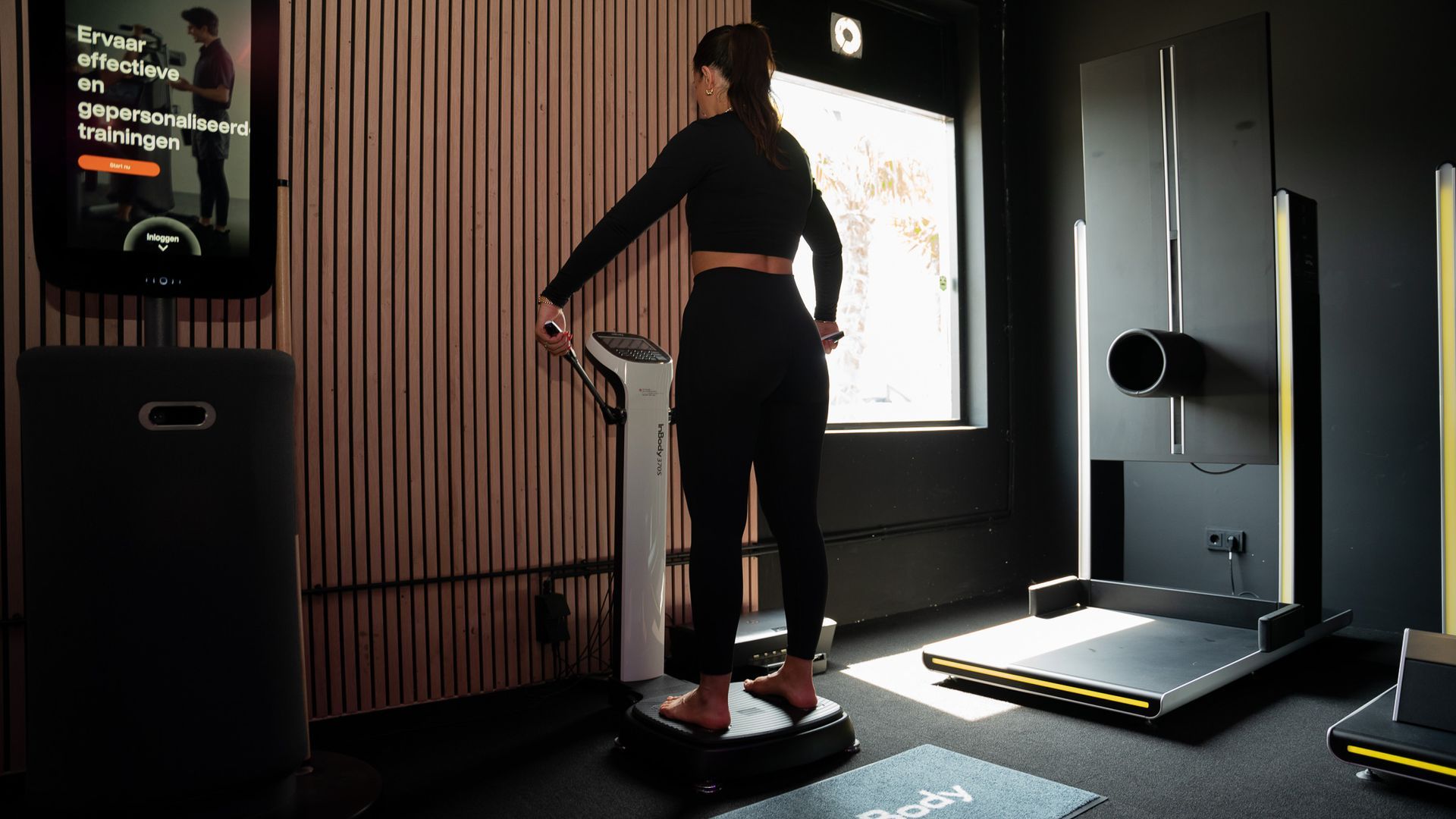 Een vrouw in zwarte sportkleding staat op een lichaamssamenstellingsweegschaal in een sportschool.