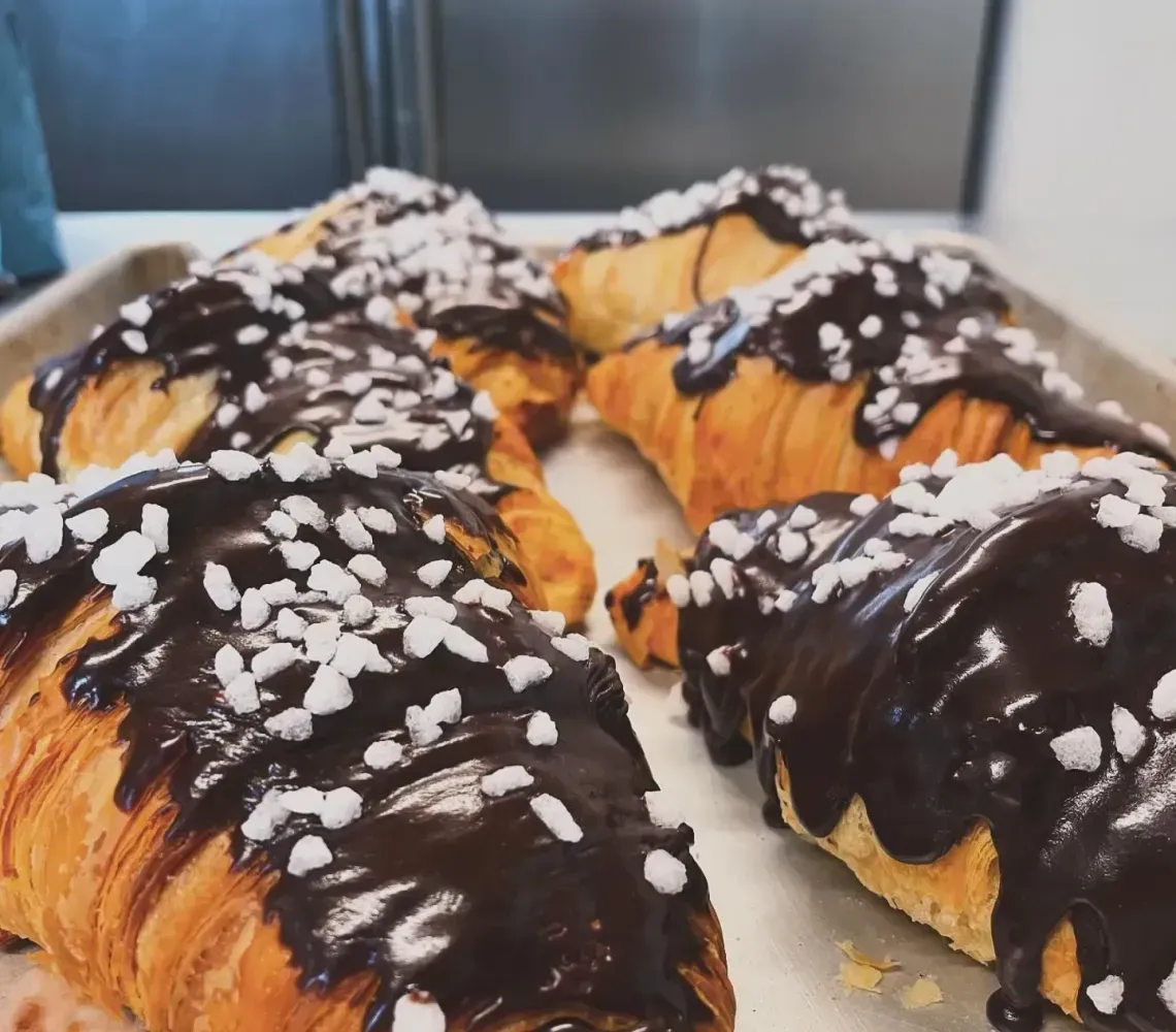 Freshly baked croissants drizzled with chocolate glaze and topped with white sugar crystals on a baking tray.