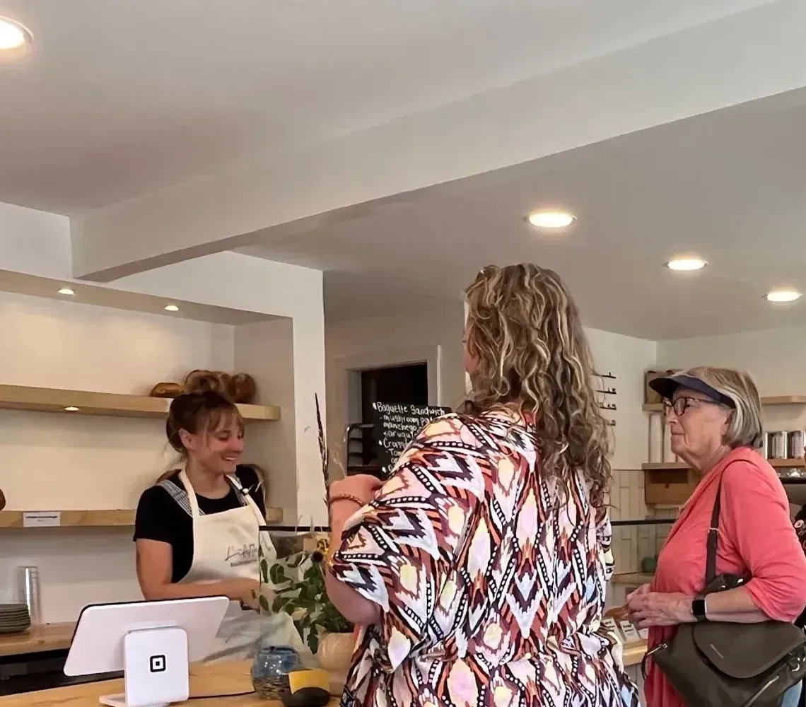 A bakery staff member stands behind a wooden counter serving customers in a bright, modern shop.