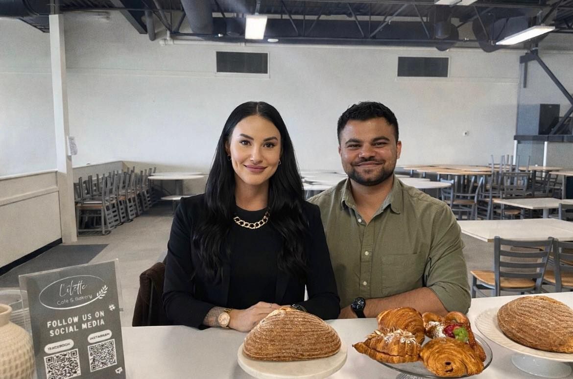 A woman and a man sit behind a table displaying various baked goods, with a sign and empty chairs in a dining area.