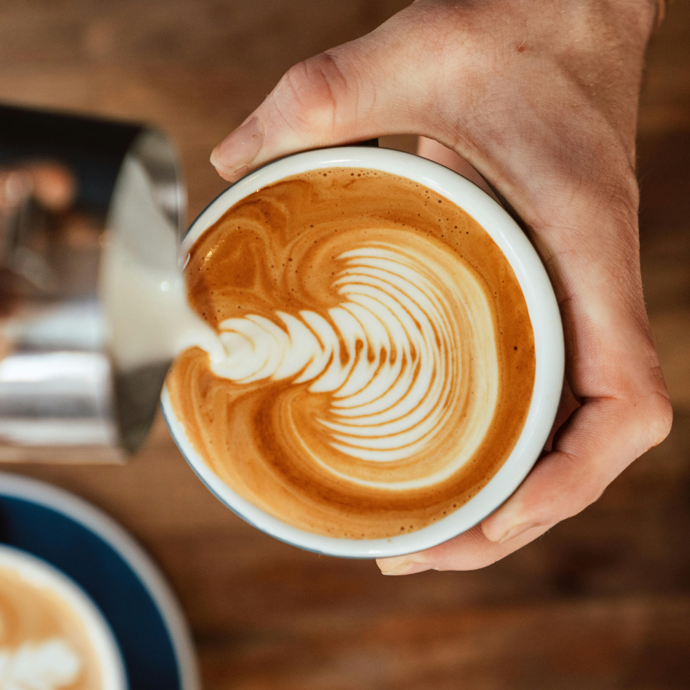 A hand pours steamed milk into a cup of espresso, creating an intricate rosetta latte art pattern.