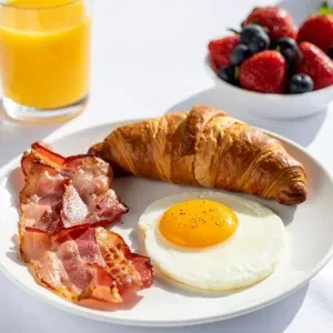 A breakfast plate with a fried egg, bacon, and a croissant, next to a bowl of berries and a glass of orange juice.