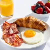 A breakfast plate with a fried egg, bacon, and a croissant, next to a bowl of berries and a glass of orange juice.