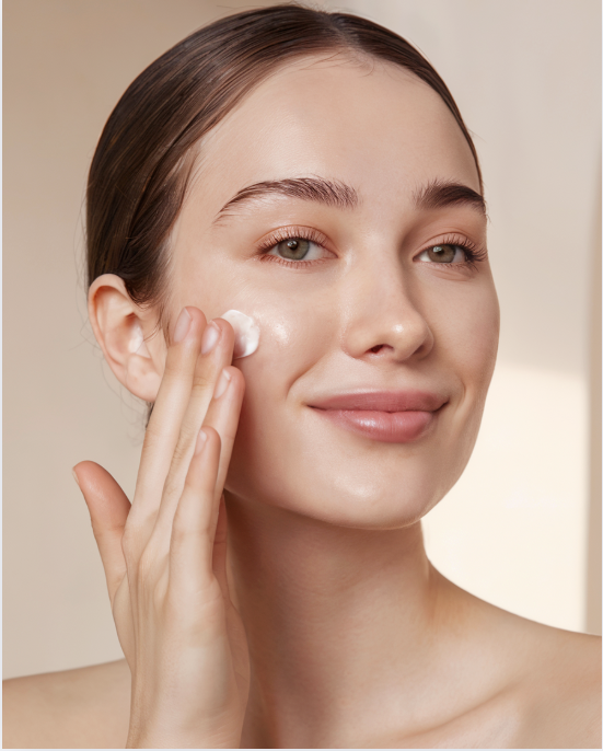 Woman applying face cream; light skin, brown hair, natural lighting, smiling, hands touching cheek.