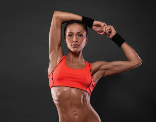 Fit woman in orange sports bra, arms raised, flexing muscles, against a gray background.