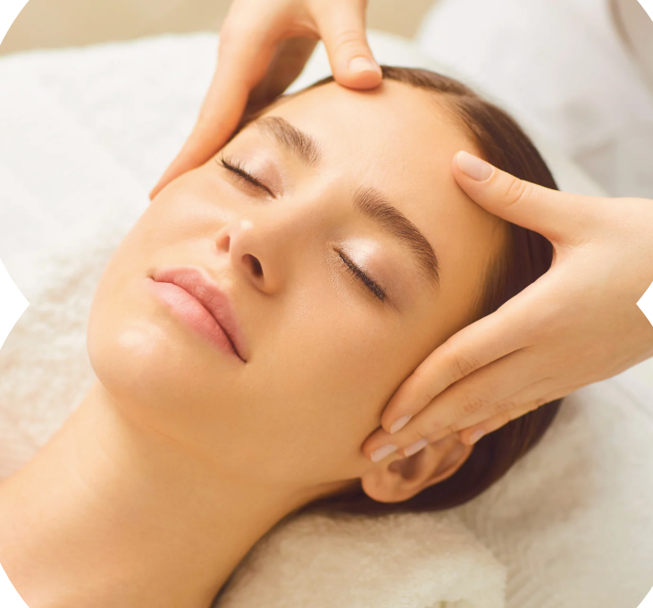 Woman receiving a facial massage, eyes closed, white setting. Hands on forehead and cheeks.