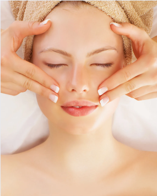 Woman receiving a facial massage with closed eyes, towel on head, hands on cheeks.