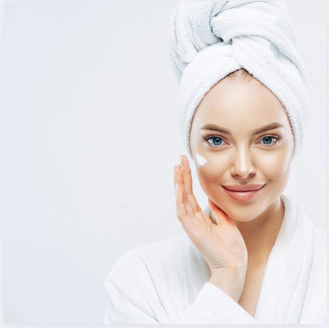 Woman in bathrobe and towel applies moisturizer to face.