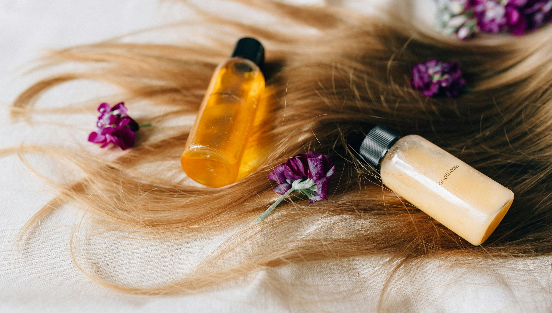 Bottles of hair product on flowing blonde hair, with purple flowers scattered around.