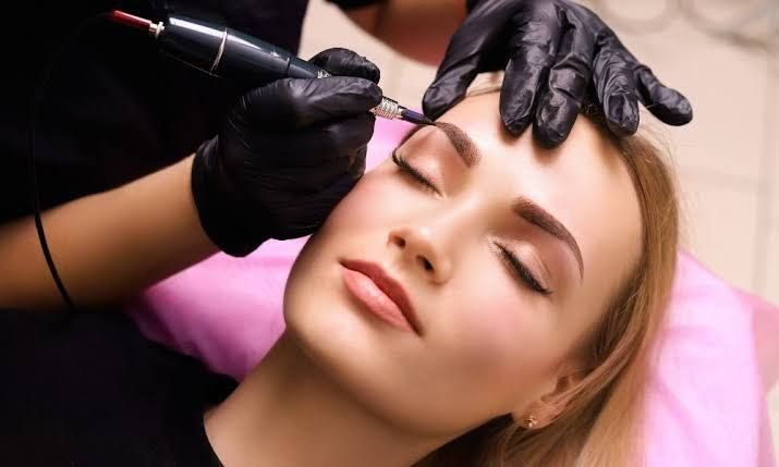 Woman receiving eyebrow microblading, artist with black gloves using a tattoo machine.