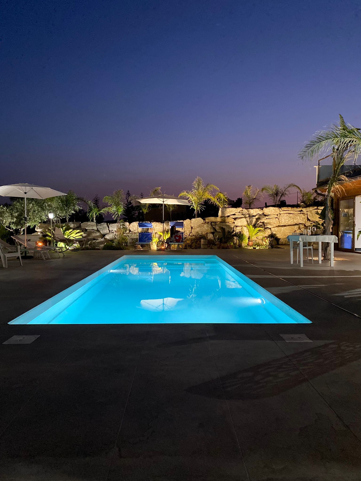 Vista notturna di una piscina rettangolare con acqua blu brillante, circondata da un patio buio e da edifici.