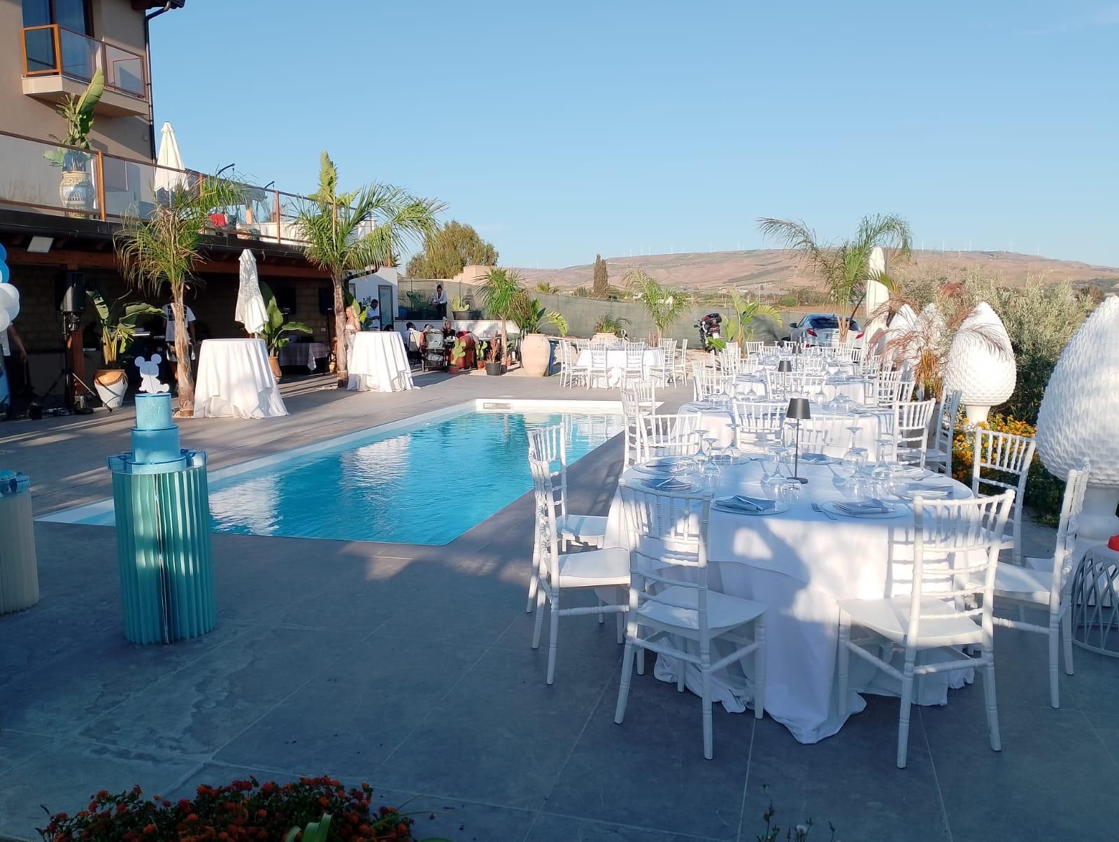 Evento all'aperto con tavoli bianchi apparecchiati per cenare vicino a una piscina sotto un cielo azzurro.