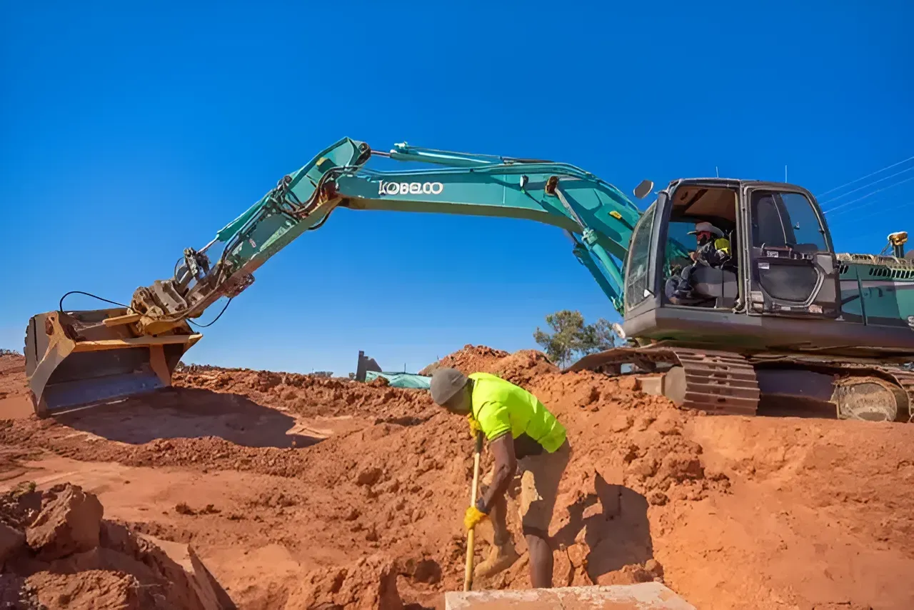 A Man is Standing in the Dirt Next to a Large Excavator — ASPLUM & CIVIL In Alice Springs, NT