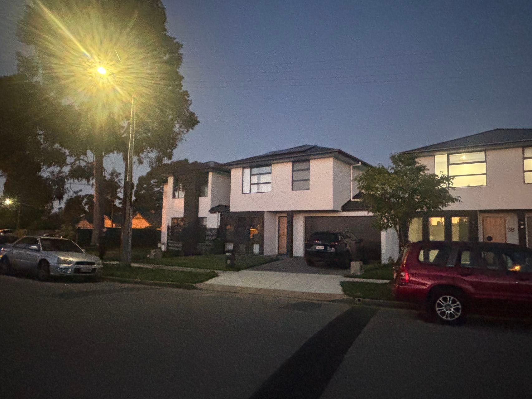 Two-story beige house with a car parked in the driveway.  Street and other cars are visible. Lit by a street lamp.