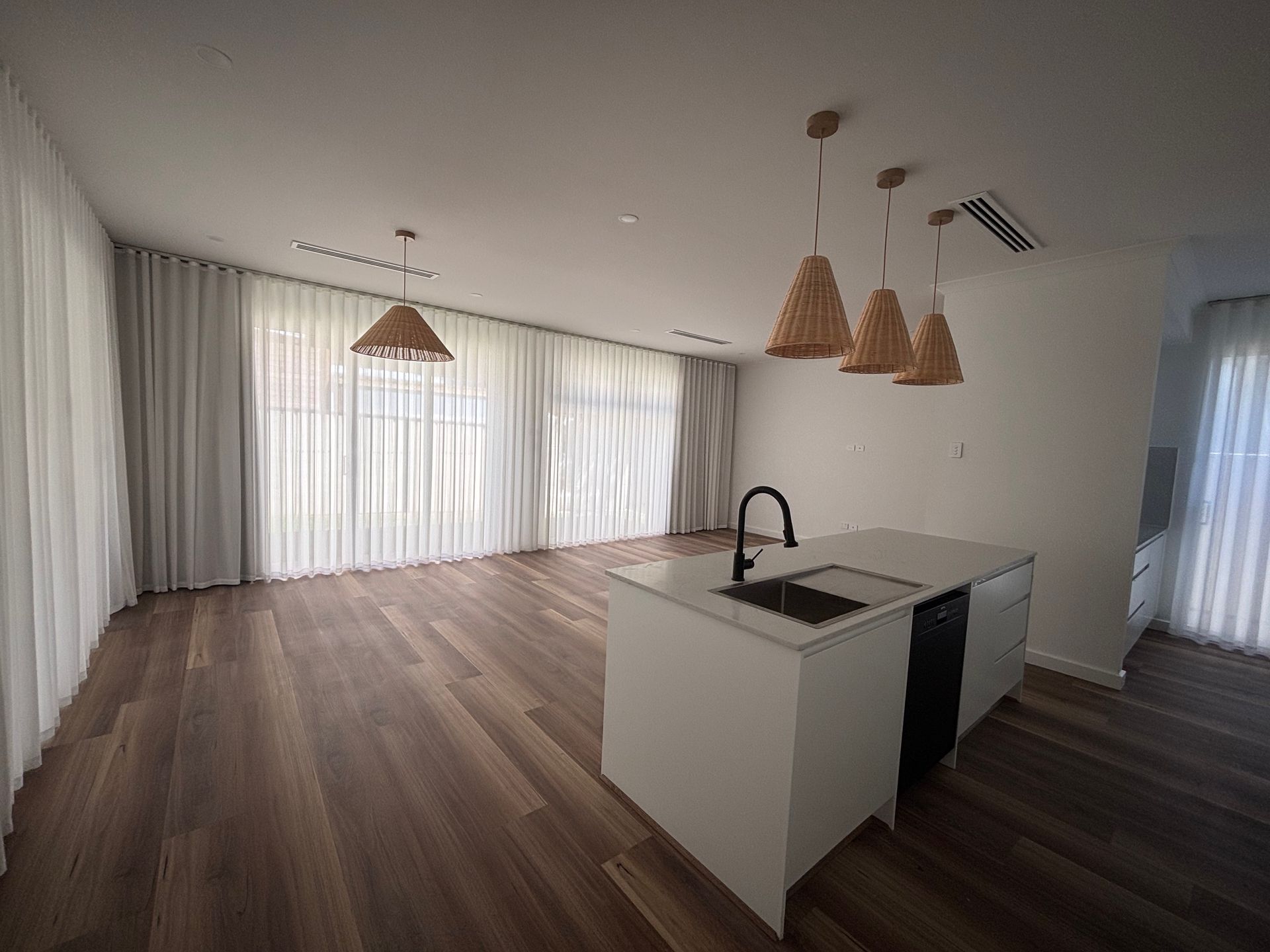 Spacious kitchen with wooden floors, white island, and woven pendant lights. Large windows with white curtains.