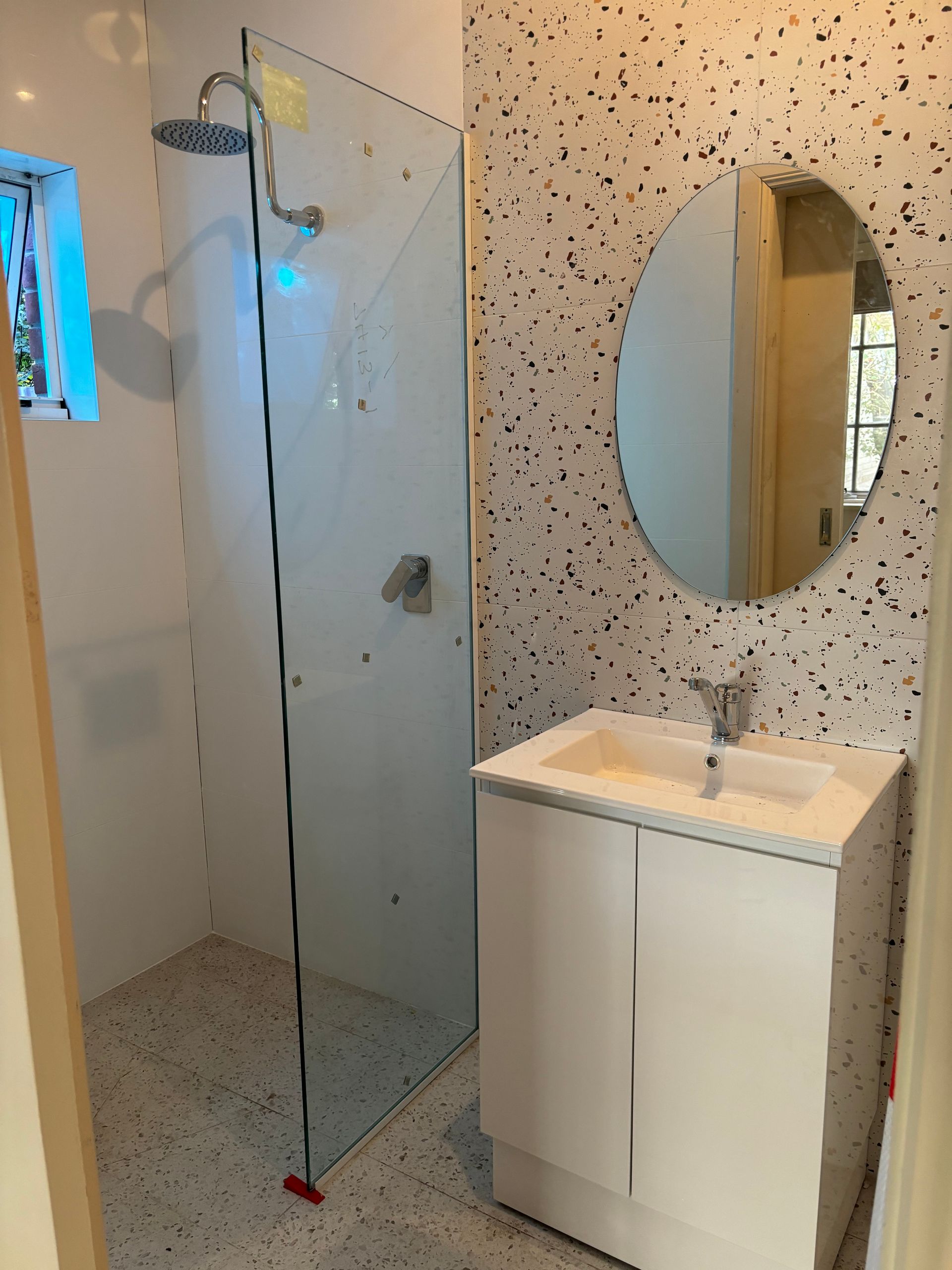 Small bathroom with a white vanity, oval mirror, and glass shower enclosure; speckled tile.