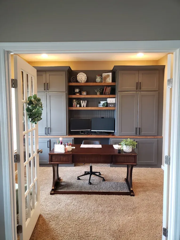 Home office with built-in gray cabinets, wooden desk, two computer monitors, and a decorative wreath.