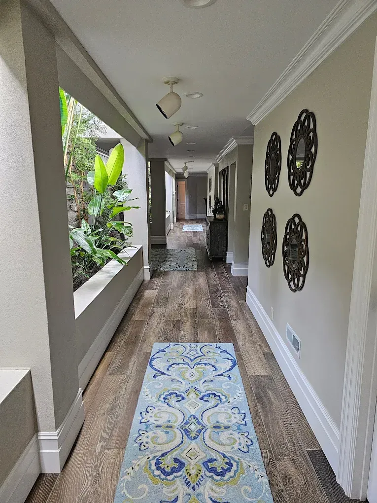 Long hallway with wood-look flooring, blue runner rug, and wall sconces. View into an outdoor garden.