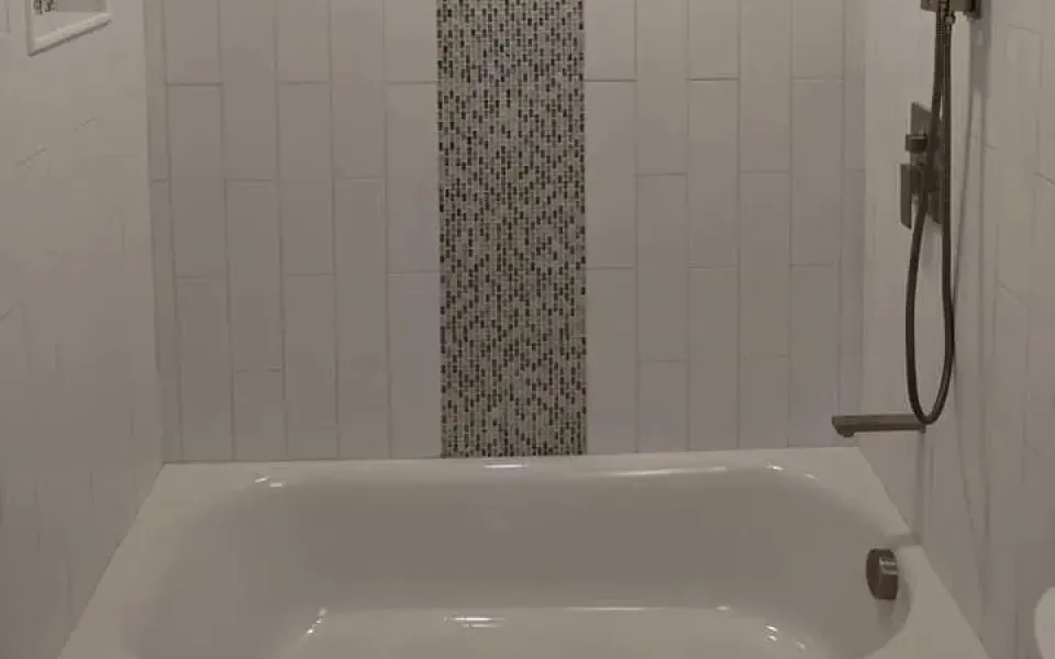 White tiled bathroom with a bathtub, patterned accent tile, and a wall-mounted showerhead on the right.
