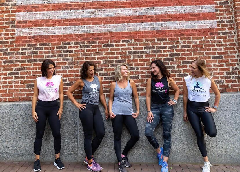 Five women in workout clothes pose in front of a brick wall with an American flag design.