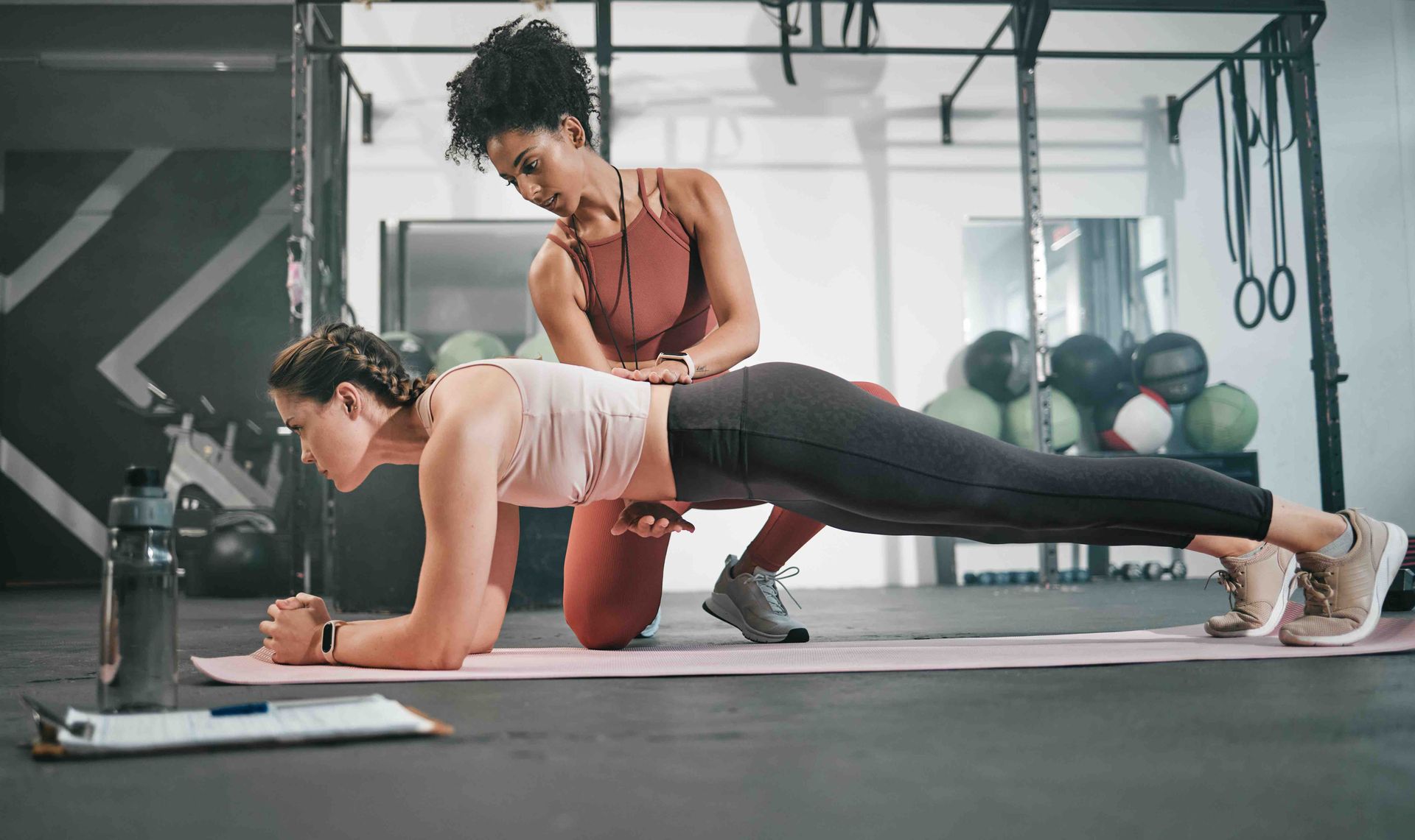 Woman in plank pose with a trainer adjusting her form in a gym. Pink mat, water bottle.