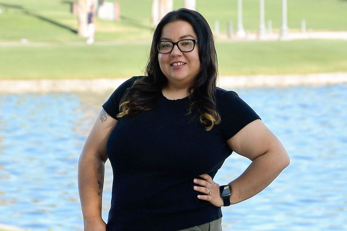 Smiling person in a black shirt standing by a pond with a grassy park in the background
