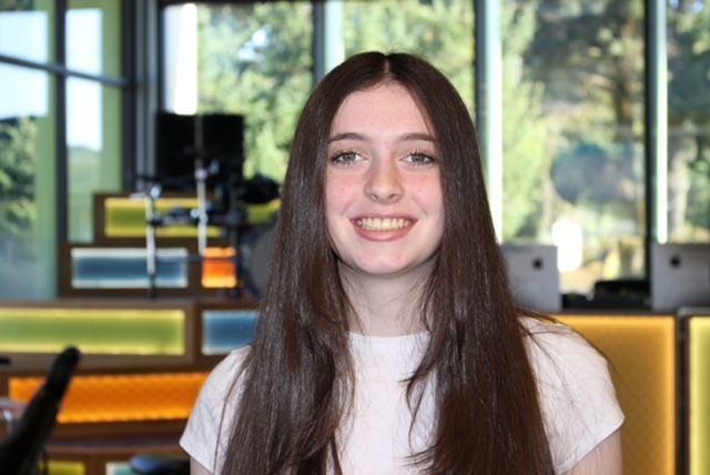 Young woman smiling, long brown hair, white top, in a brightly lit studio with green backdrop.