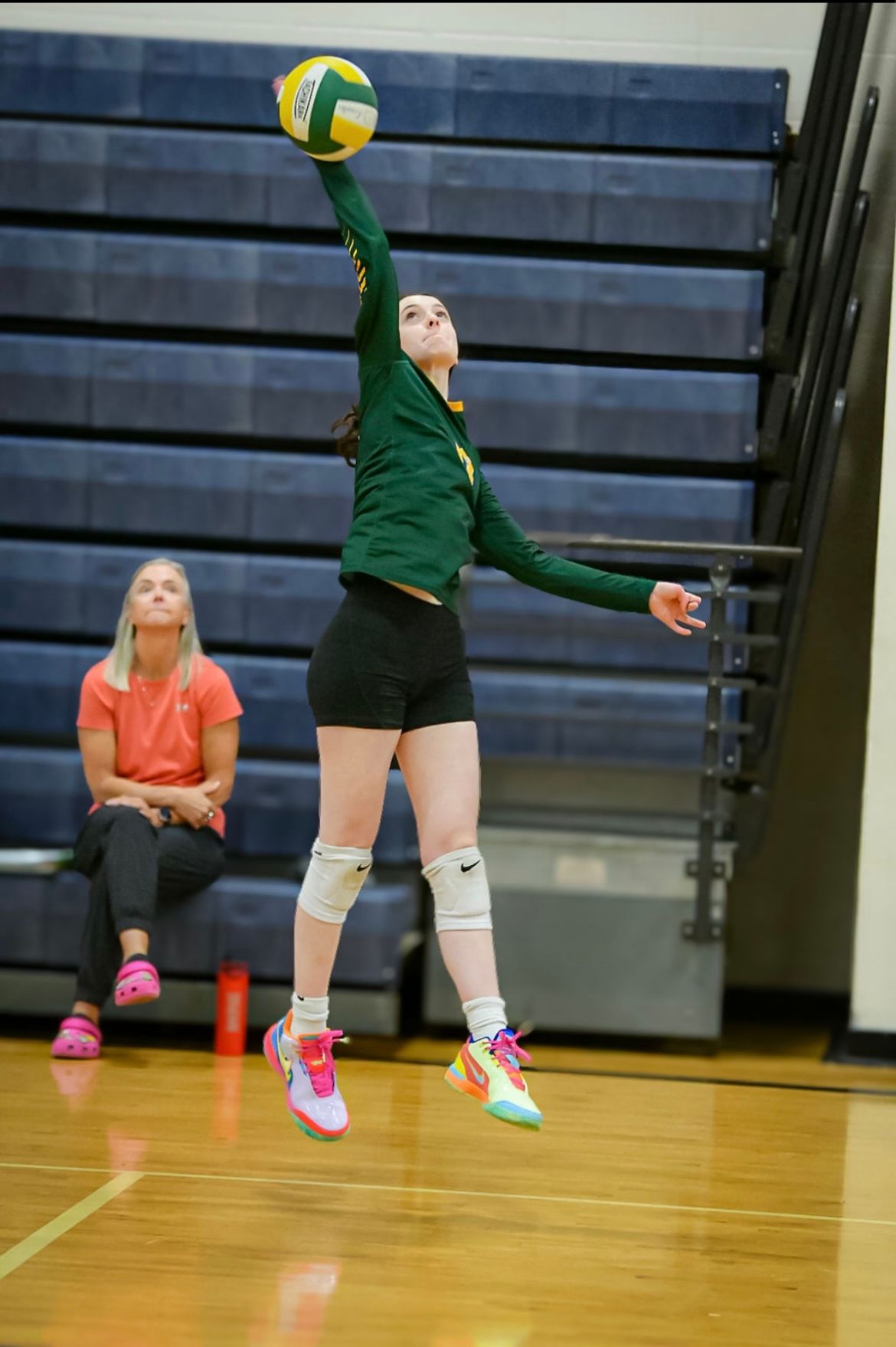Volleyball player in green and black jumping to spike. Another person watches.