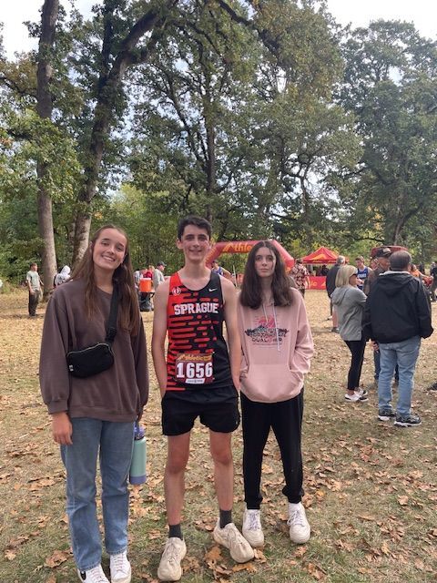 Three people posing outdoors; cross-country runner in race bib stands between two friends; autumn setting.