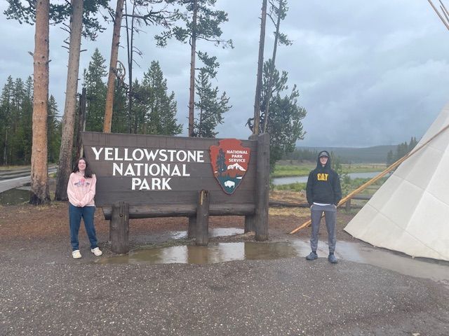 Two people pose near Yellowstone National Park sign under a cloudy sky; a teepee is visible to the right.