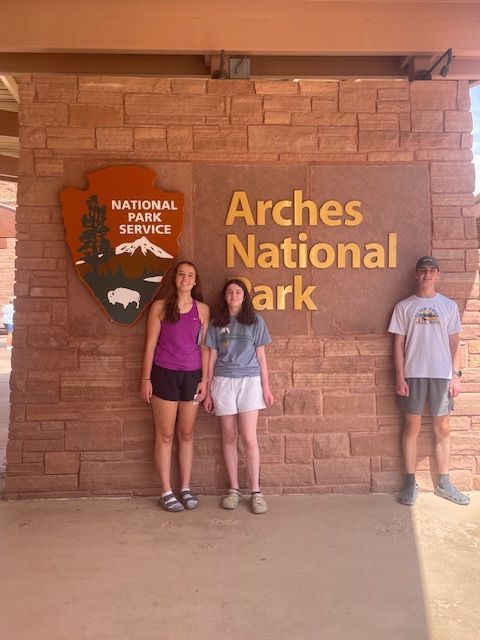 Three people stand in front of the Arches National Park sign.