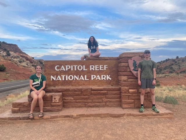 Three people pose by a Capitol Reef National Park sign, against a backdrop of red rocks and cloudy sky.