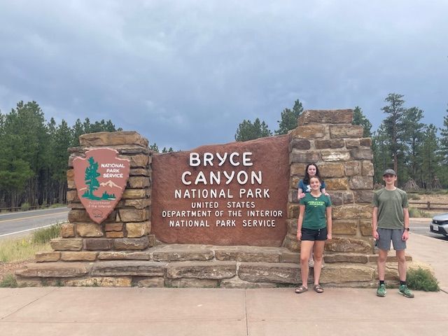 Three people pose in front of the Bryce Canyon National Park sign. Overcast day.