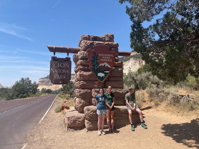 Three people pose near the Zion National Park entrance sign on a sunny day.