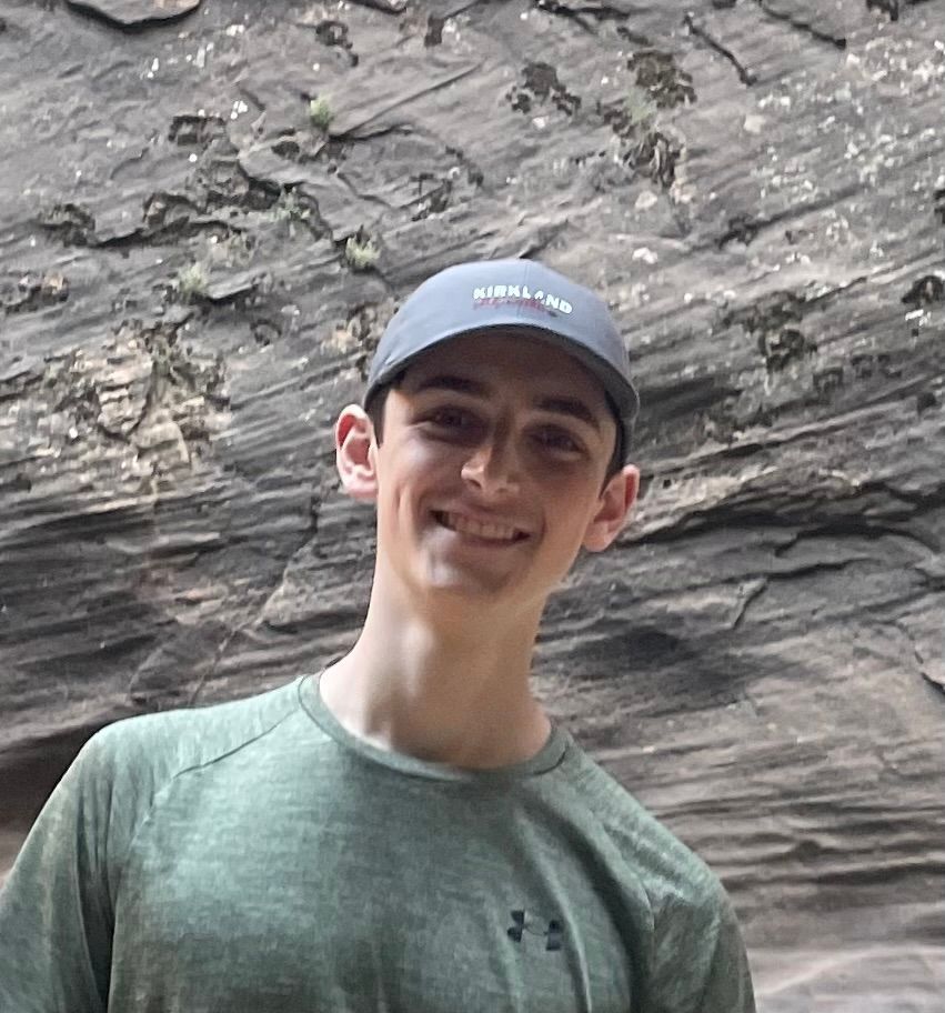 Young man smiles, wearing a cap and green shirt, standing against a textured rock background.