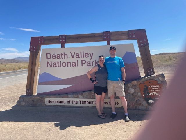 Two people pose in front of a Death Valley National Park sign under a blue sky.