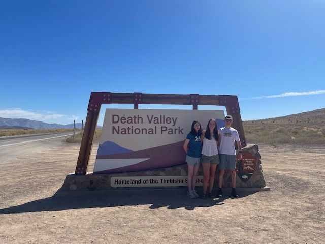Three people stand in front of the Death Valley National Park sign on a sunny day.