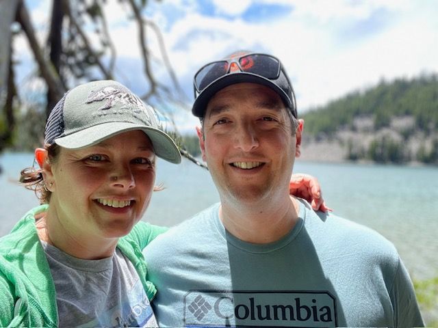 Couple smiling, posing outdoors by a lake, wearing hats and casual clothing.