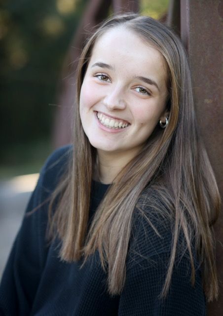 Young woman with long brown hair smiles broadly outdoors, leaning against a rusty bridge.