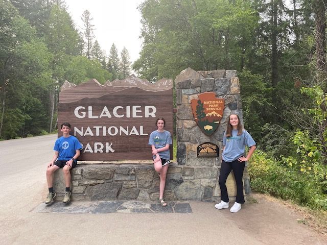 Three people pose by the Glacier National Park sign, a wooden brown sign with stone base, surrounded by trees.