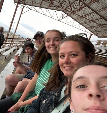 Group of people smiling, sitting in outdoor stadium seating. Cloudy sky above.