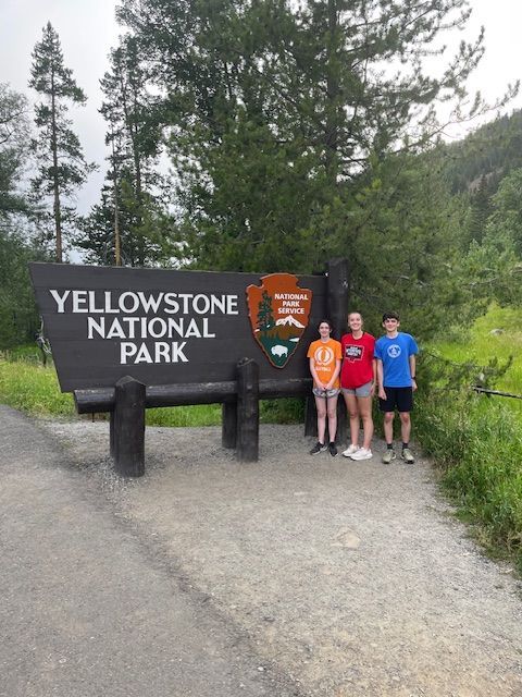 Three people pose in front of the Yellowstone National Park sign on a paved path, with trees in the background.