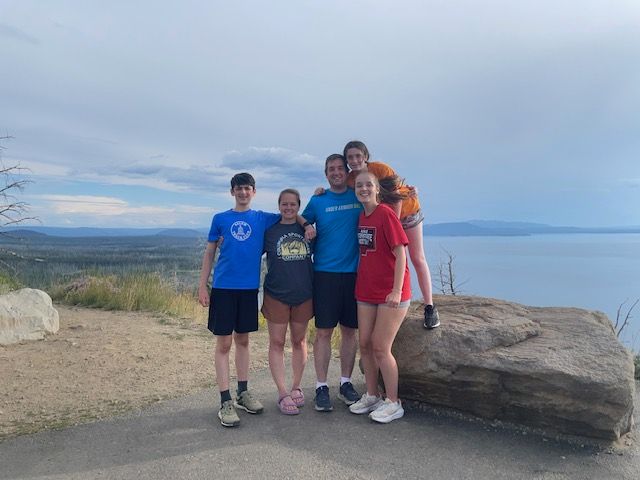 Family of five smiles, posing on a rock overlooking a lake and landscape under a cloudy sky.