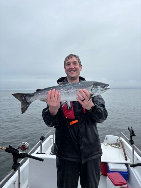 Man on a boat holding a silver salmon, smiling. Cloudy sky, gray water.