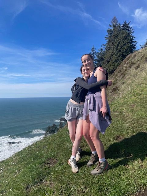 Two women hugging on a grassy cliff overlooking the ocean under a blue sky.