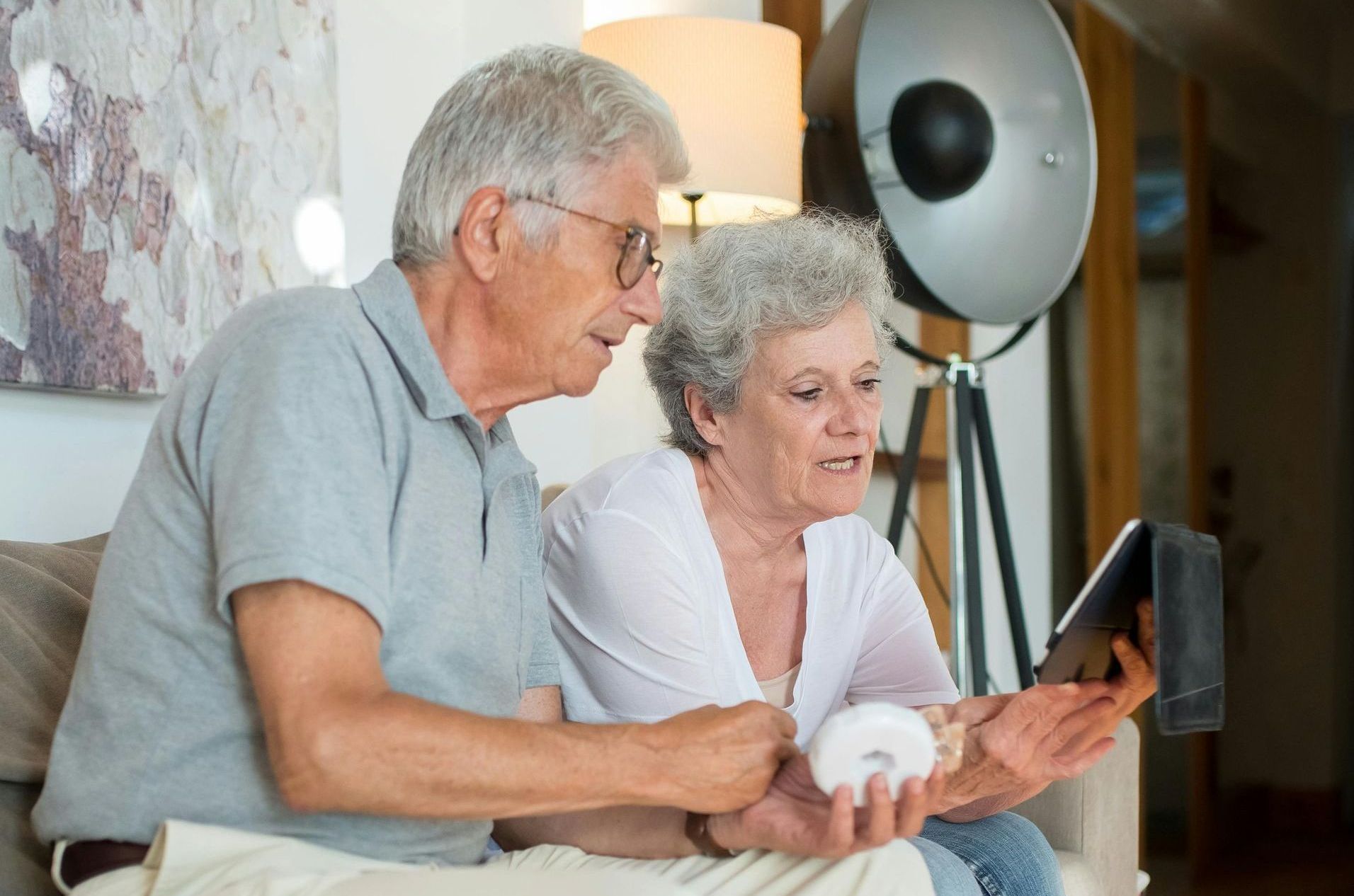 Elderly couple looking at tablet, smiling, indoors. Man wearing glasses, gray hair, woman holding donut.
