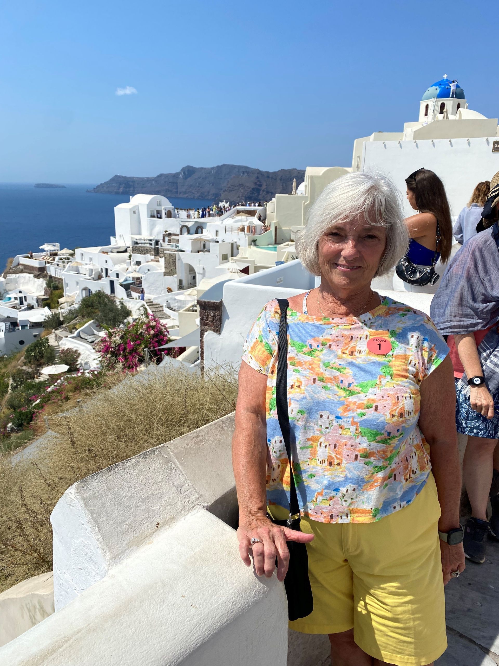 A woman in a floral shirt and yellow shorts is standing on a balcony overlooking the ocean.