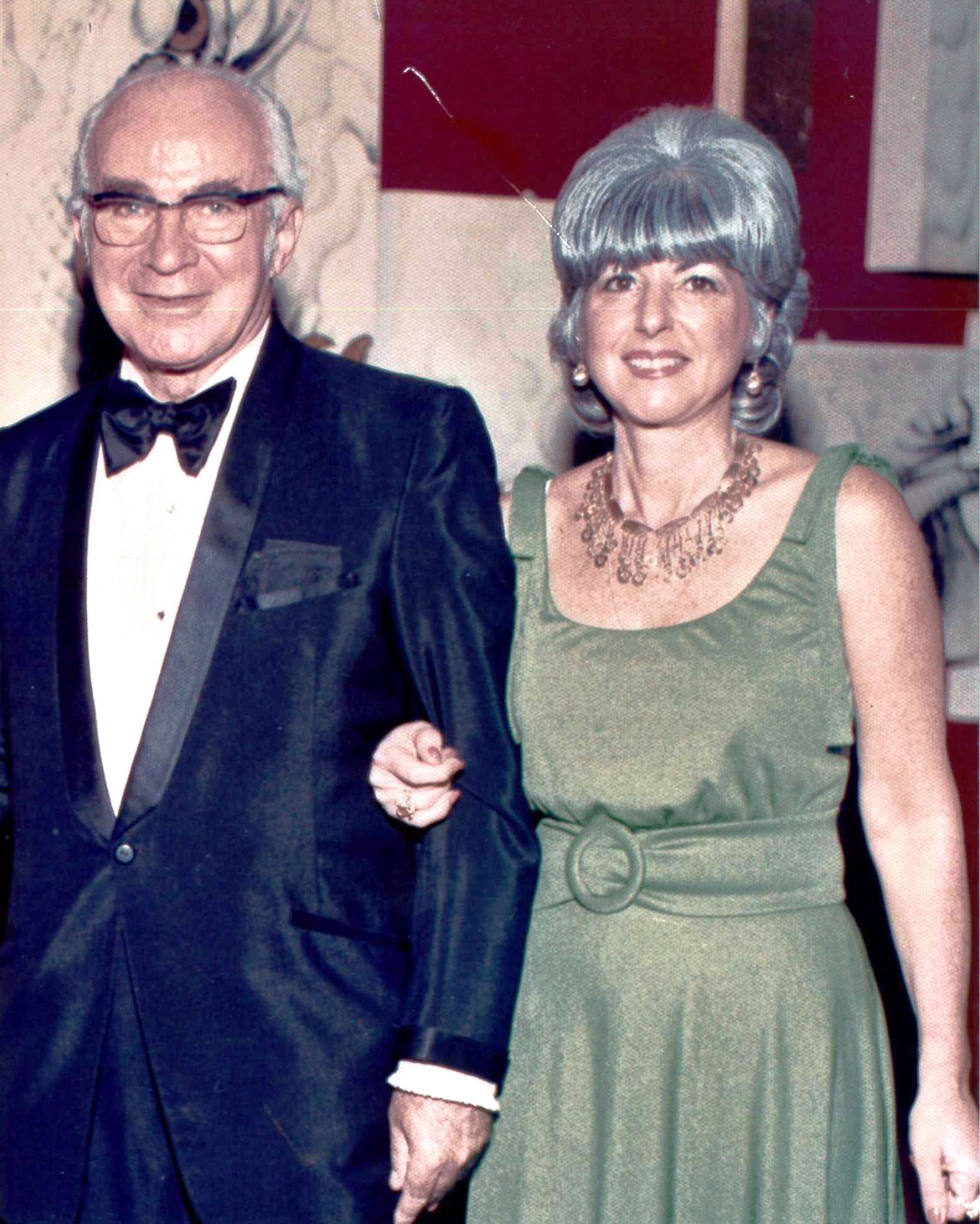 A man in a tuxedo and a woman in a green dress are holding hands