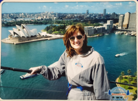 A woman stands on a balcony overlooking a body of water with the opera house in the background