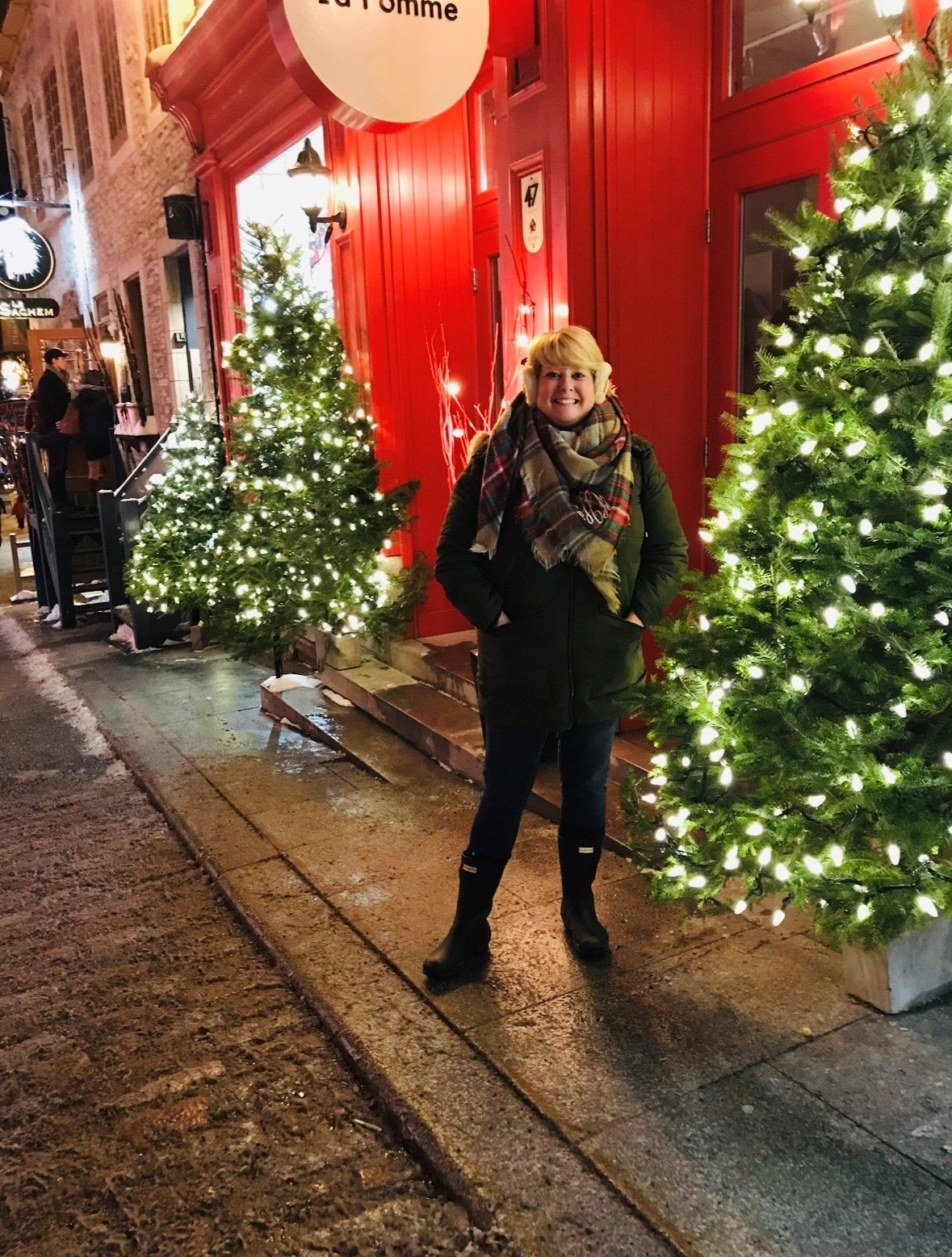 A woman is standing in front of a red building with christmas trees in front of it.